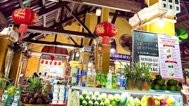 A vibrant market stall resembles a lively cafe, displaying fresh fruits, juice, and colorful lanterns under a wooden roof.