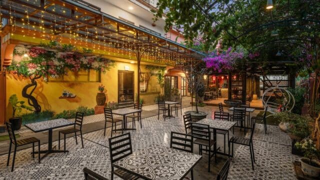Charming outdoor café with patterned tile floor, black tables, string lights, and lush greenery under a canopy.