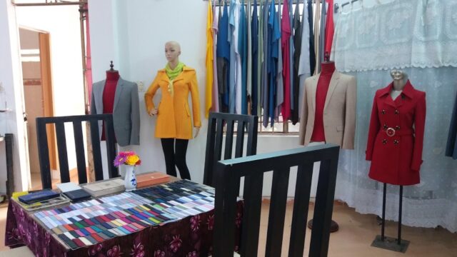 Hoi An tailoring shop with mannequins in coats, fabric samples on a table, and clothes hanging by the window.