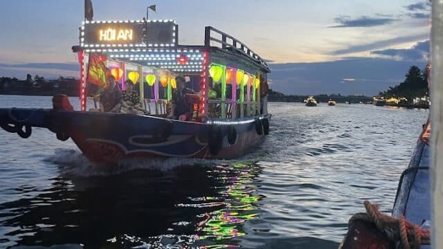A colorful, illuminated boat labeled Hoi An floats on a river at dusk, reflecting lights in the water—an iconic view with Onion Phi Tours.