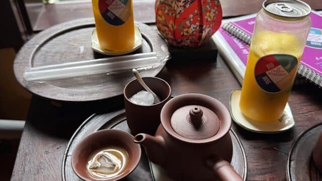 Tea set and two iced drinks on a wooden table at Ceramics Cafe, with a red decorative item and notebook nearby.