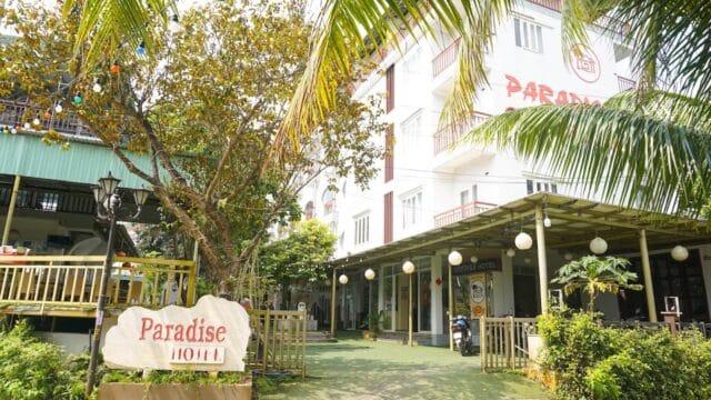 Paradise Hotel building with a large sign offers luxury accommodation amid greenery and palm trees in a sunny outdoor setting.