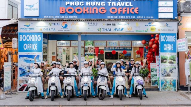 Nine people in matching outfits and helmets sit on scooters outside a brightly colored Hoi An travel booking office.