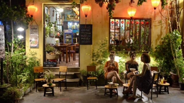 Three women sit outside a cozy, lantern-lit café at night, surrounded by plants and warm yellow lights.