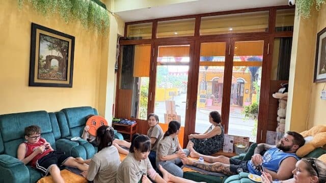 People relaxing and getting foot massages at Potri Nails & Spa in a cozy, brightly-lit room with green sofas and hanging plants.