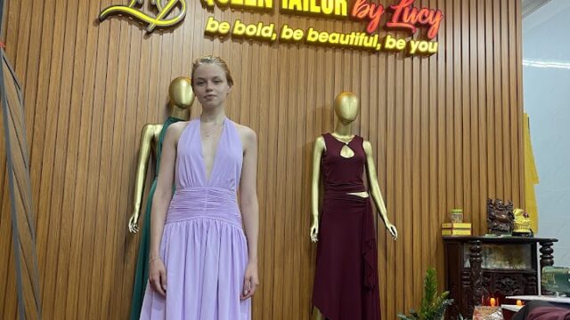 A woman in a lilac halter dress stands in a tailor shop with mannequins and a Queen Tailor by Lucy sign.