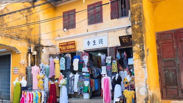 A yellow building with lanterns and clothes displayed outside a shop on a charming Hoi An old town street.