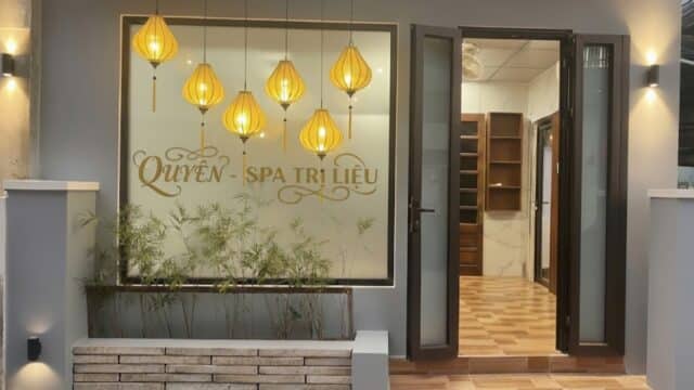 Front view of a spa with warm hanging lights, glass door open, and QUYEN - SPA TRI LIEU on the window.