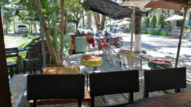 Outdoor cafe in Hoi An with colorful tire seats, tables, and shade, overlooking a street with trees and parked scooters.