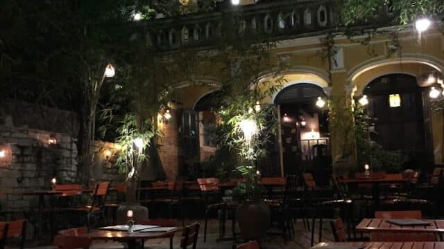Riverside restaurant at night with empty wooden tables, soft lights, and a large tree glowing with green lighting.