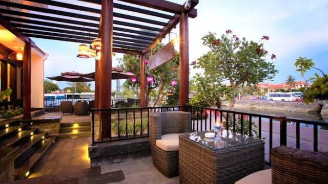 Outdoor patio at River Suites Hoi An with wicker chairs, glass table, lush plants, lanterns, and evening lights near the street.