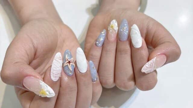 Hands with long, almond-shaped nails featuring blue, white, and pink designs with starfish and seashell accents at Rosan De Beauty spa.