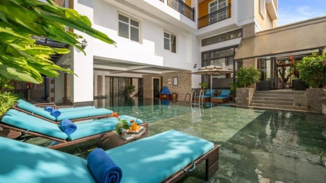 Turquoise lounge chairs by a clear pool at Royal Riverside Hotel, beside a modern building with greenery and sunlight.