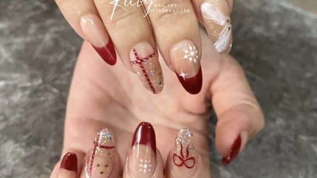 Hands with festive nail art: red French tips, glitter, bows, and holiday-themed designs on a gray textured background.