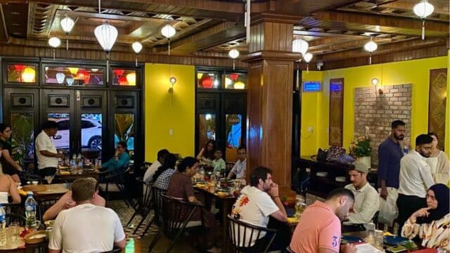 Busy Indian Restaurant with diners at tables, saffron-hued walls, wooden decor, and glowing lantern lights.