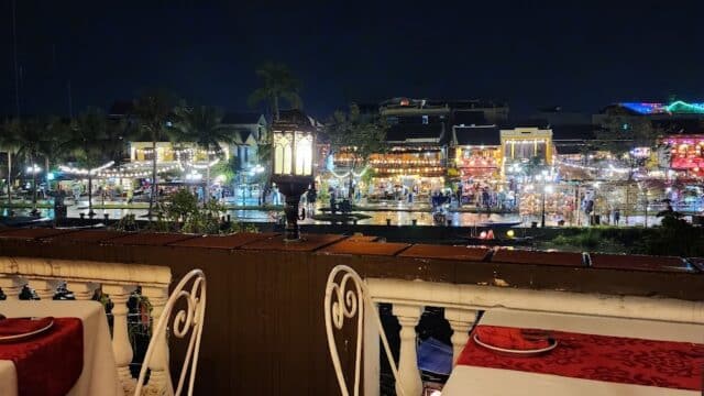 A balcony restaurant at night overlooks a lively, brightly lit riverside area with palm trees.