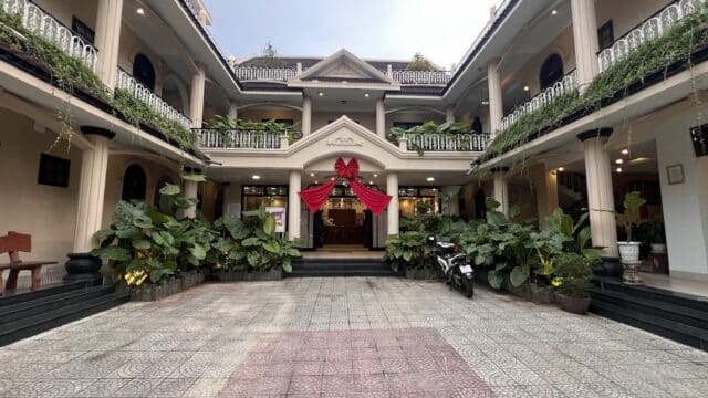 Two-story Sala Hoi An Hotel with plants, columns, and a large red bow above the entrance in a courtyard.