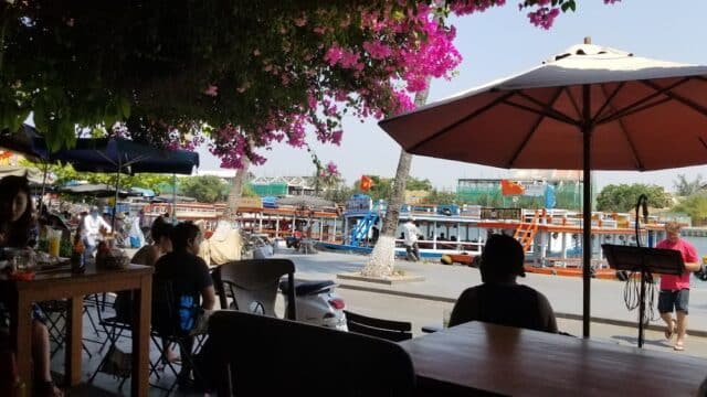 Outdoor café at Sao Mai Restaurant, with people under umbrellas, pink flowers overhead, and boats docked by the riverside.