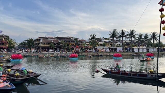 Colorful boats and large pink lanterns float on a riverside in lively Hoi An, with palm trees, buildings, and SEA Tours Hoi An nearby.