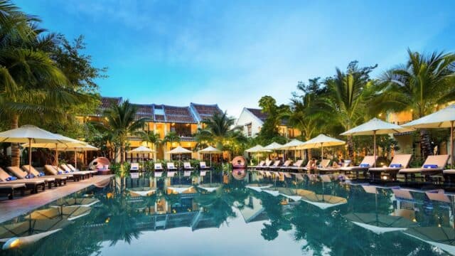 Luxury resort pool surrounded by lounge chairs, umbrellas, palm trees, and Silk Village-inspired tropical buildings at sunset.