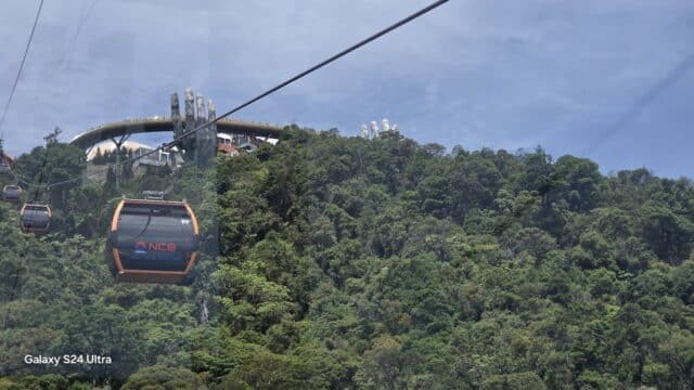 Cable cars travel over a lush green forest in Vietnam Travel, with a bridge held by giant stone hands under a clear blue sky.
