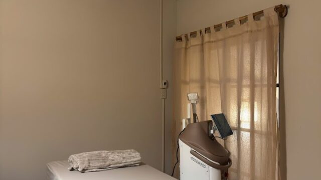 A treatment room with a covered massage table, medical device, beige curtains, and an air conditioner.