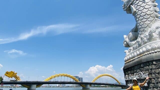 A woman in a yellow dress poses by a dragon statue near Hoi An’s yellow dragon bridge over a river under a blue sky.