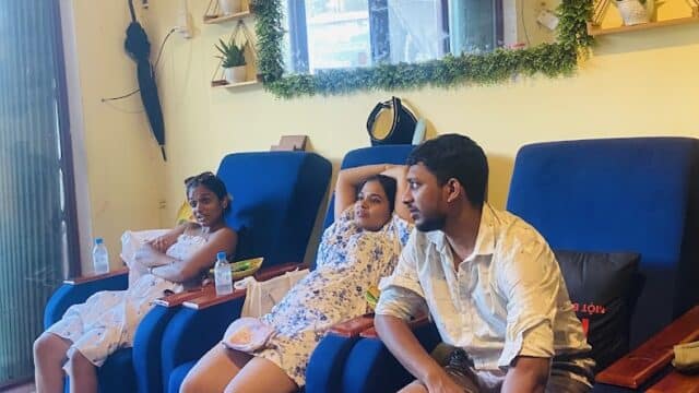 Three people relaxing in blue chairs at a brightly lit spa or salon with yellow walls and decorative plants.