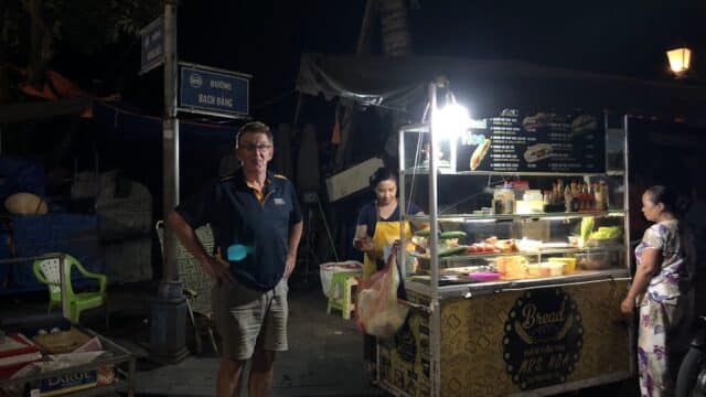 A man stands by a brightly lit street food cart at night in Hoi An, with two women preparing food behind the counter.