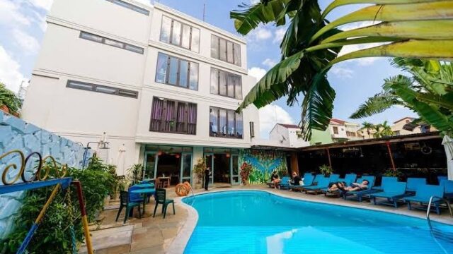 Modern Sunflower Hotel with a swimming pool, blue lounge chairs, and people relaxing under palm trees on a sunny day.