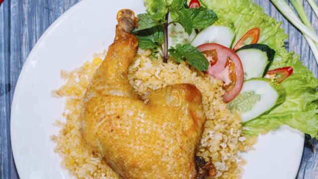 A plate of Hoi An fried chicken leg on yellow rice, served with lettuce, cucumber, tomato, and mint garnish.