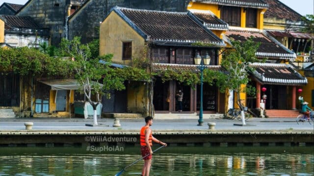 A man paddleboards on a river in Hoi An, gliding past yellow historic buildings in this charming Vietnamese town.