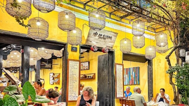 People sit outside a vibrant yellow café in Hoi An at night, surrounded by hanging lanterns and lush plants.