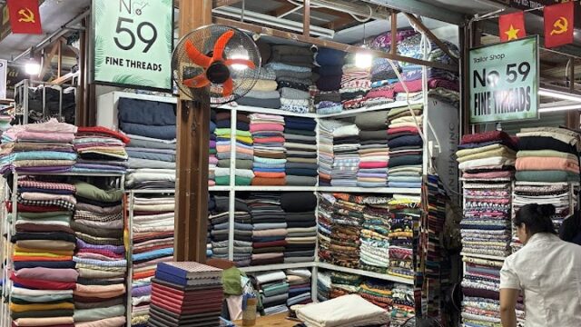 A fabric shop with stacked cloth, red chairs, and Vietnam flags and signs overhead in a market setting.