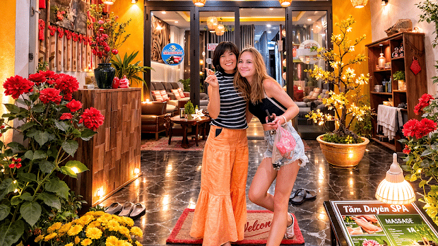 Two women smiling and posing at the entrance of Tam Duyen Spa, surrounded by flowers and warm lights.