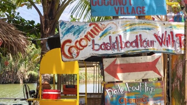 Colorful signs for Tamcoconut Basket Boat Tour by a river, with a red scooter and yellow booth under palm trees.
