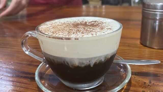A glass cup of coffee with creamy foam sits on a wooden table at Tasting Cafe, a spoon resting beside the saucer.