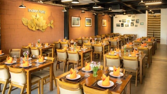 Spacious restaurant with neatly set tables, orange napkins, wall art, and Indian Tasty sign on the wall.
