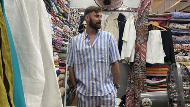 A man in blue and white striped shirt and shorts stands in a colorful Hoi An fabric store, looking to the side.