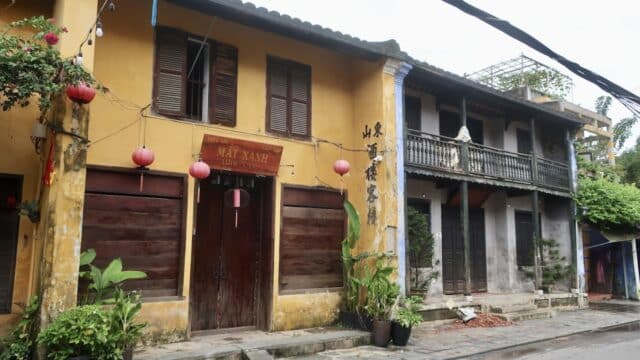 Two old yellow buildings with wooden shutters, red lanterns, and potted plants line a quiet street in Hoi An.