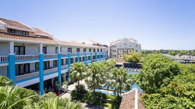 A sunny view of Thanh Binh Riverside Hotel, with blue accents, a pool, palm trees, and nearby buildings under a clear sky.