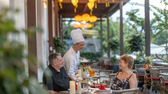 A chef serves dishes to two smiling diners at an outdoor restaurant decorated with hanging lanterns.