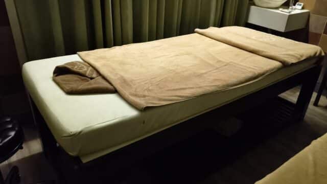 A Ginger Spa massage table with brown towels in a dimly lit room, green curtains, and a sink in the background for ultimate wellness.