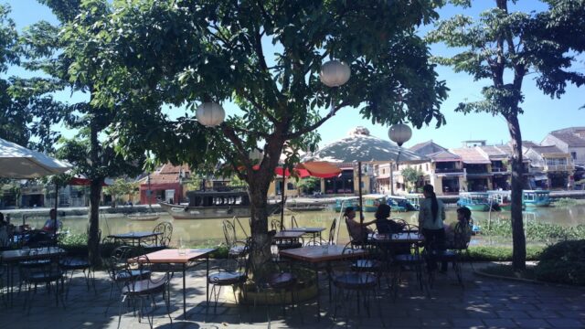 Outdoor riverside café with trees and lanterns—an Island Restaurant where people relax under umbrellas on a sunny day.