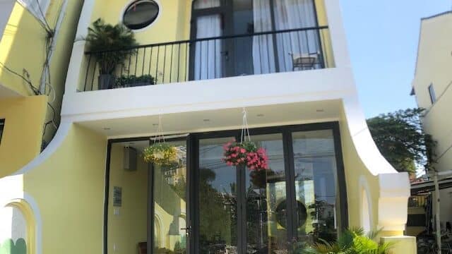 A modern yellow three-story Night Market Villa with balconies, potted plants, and a green motorcycle parked in front.