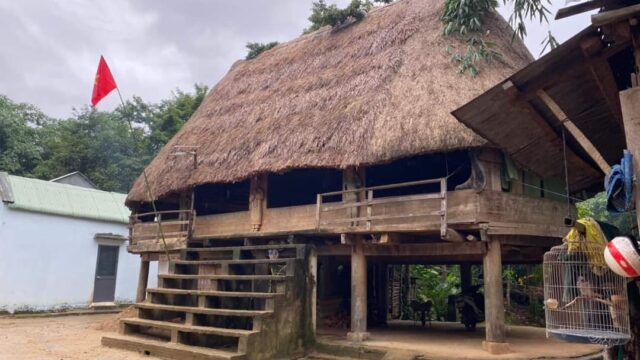 A traditional stilt house with a thatched roof and steps, a red flag nearby, under a cloudy sky—perfect for a Top Gear Tour snapshot.