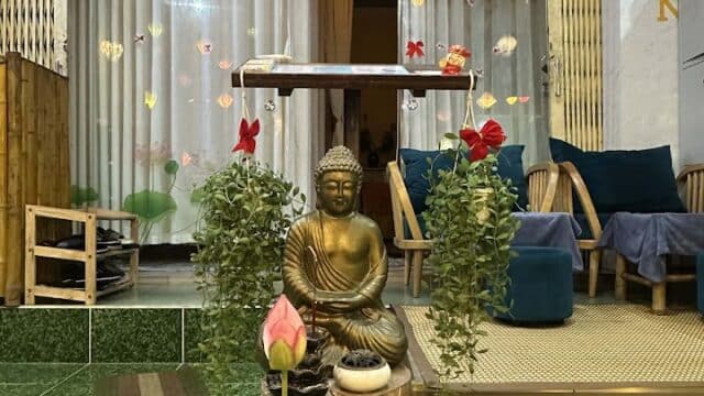 A spa entrance with a Buddha statue, plants, and a lotus flower in a pot; cozy seating in the background.