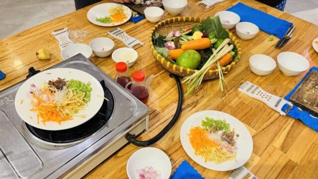 A table set for cooking in Hoi An with plates of chopped vegetables, sauces, and kitchen tools around a basket of fresh produce.