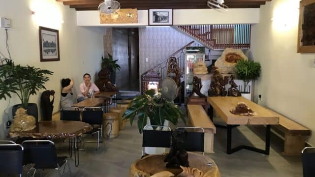 A cozy cafe with wooden tables, carved sculptures, plants, and two people sitting near the back wall.