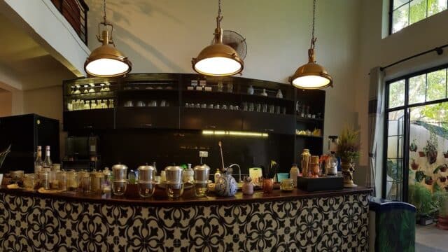 A cozy café interior in Hoi An with a patterned counter, hanging lights, and large windows overlooking a lush garden.